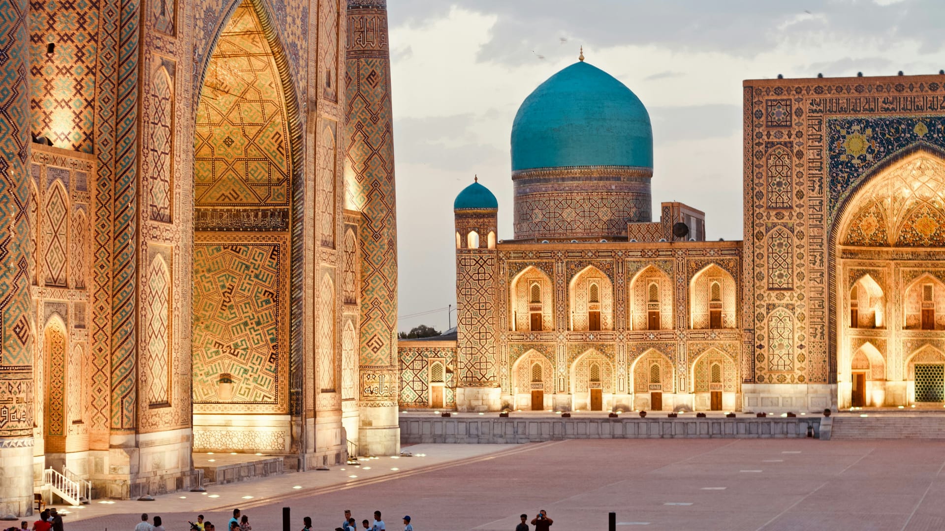 A wide panoramic view of the three madrasahs of Registan Square illuminated with warm golden lights at night.