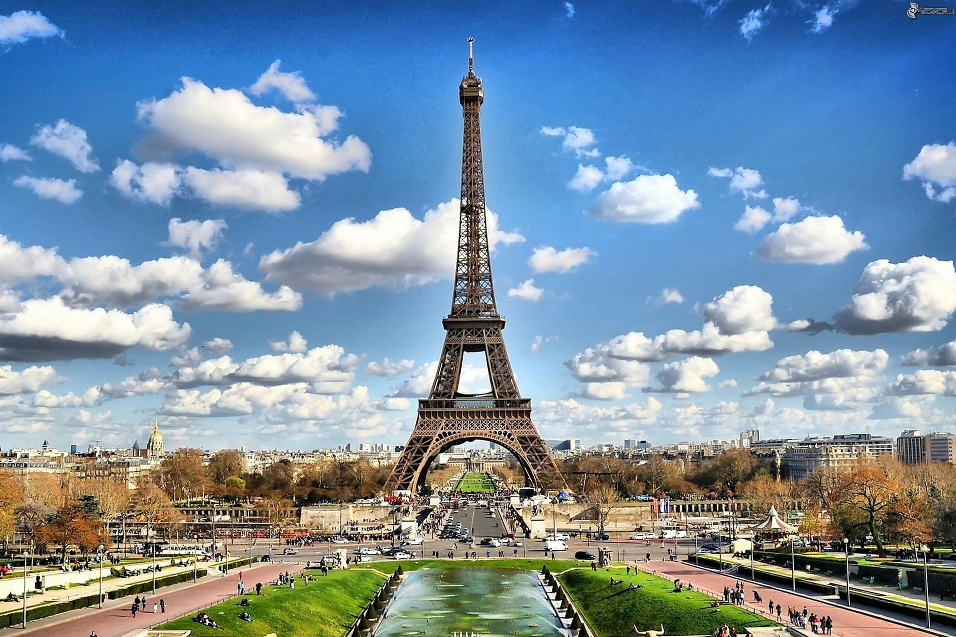 The Eiffel Tower rising into a blue sky with white clouds, viewed from the grassy lawns of the Champ de Mars.
