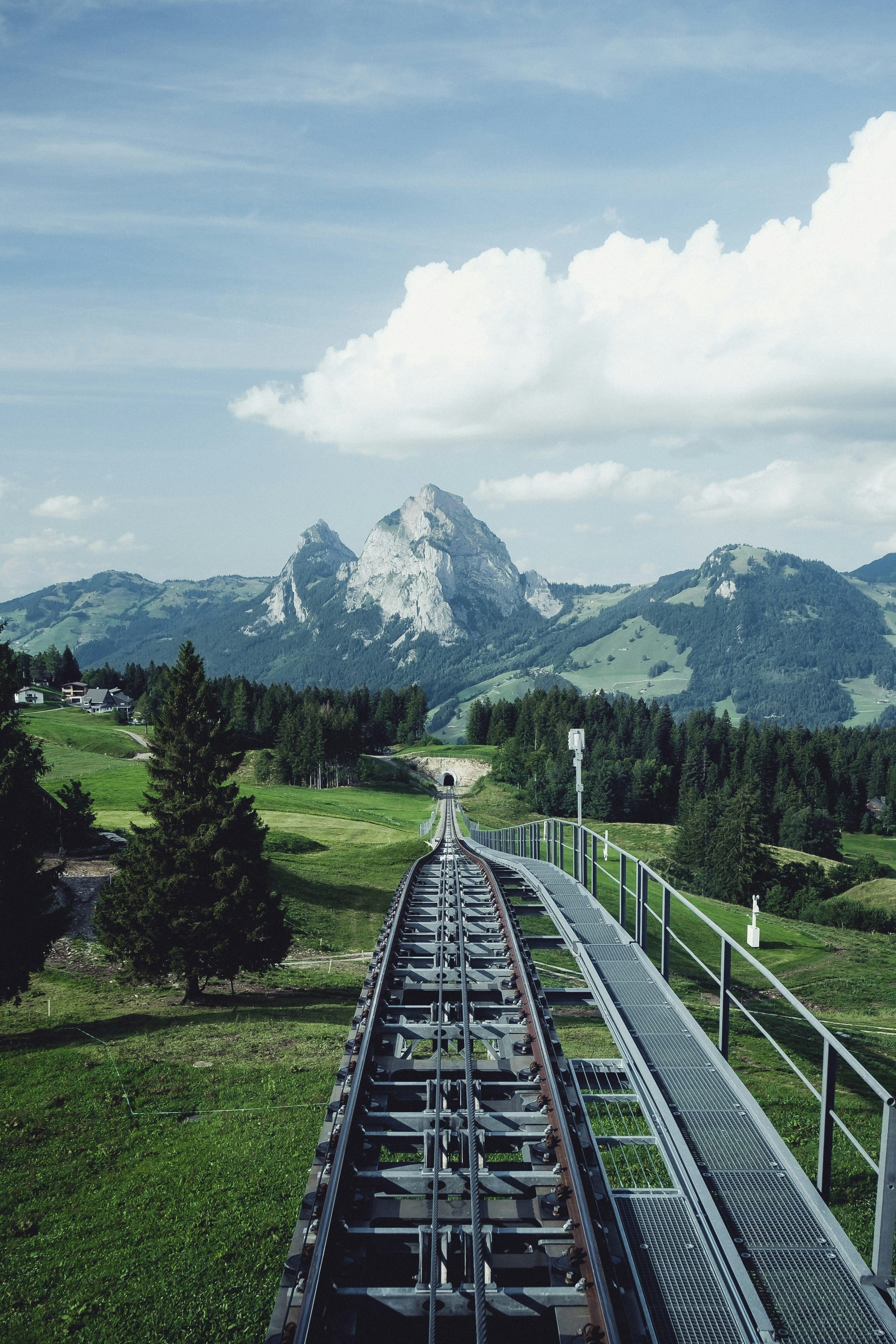 A steep funicular railway track ascending a green mountain slope towards a jagged rocky peak in the distance.