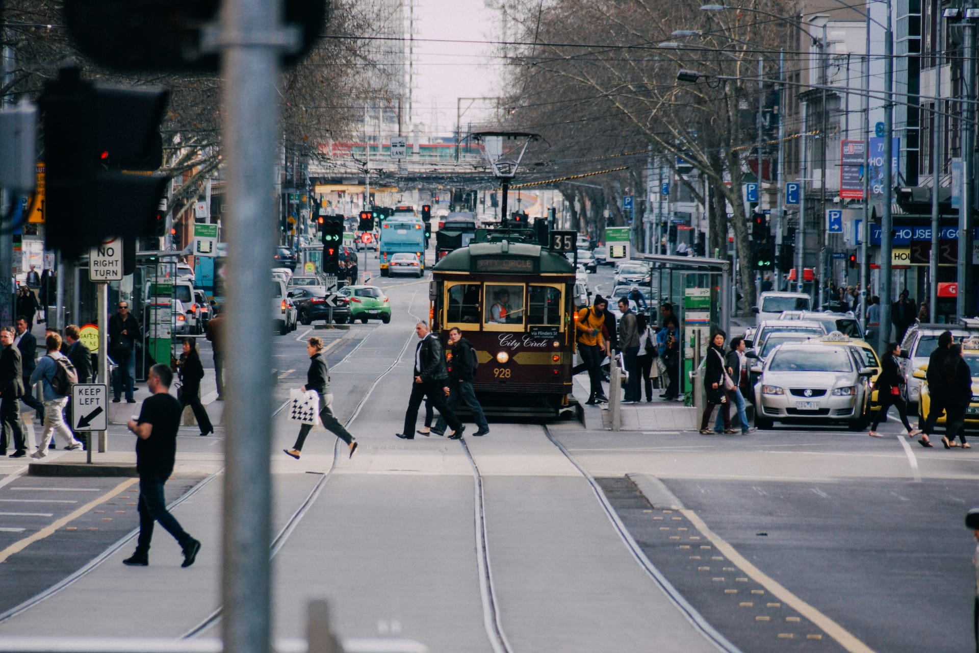 A vintage tram traveling down a busy street in Melbourne lined with trees and pedestrians