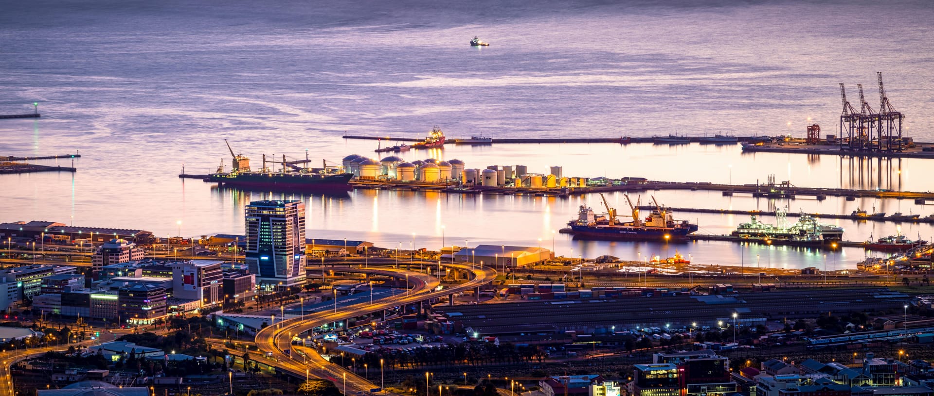 A panoramic view of the Cape Town commercial port and harbor lit up at night with city lights reflecting on the water.