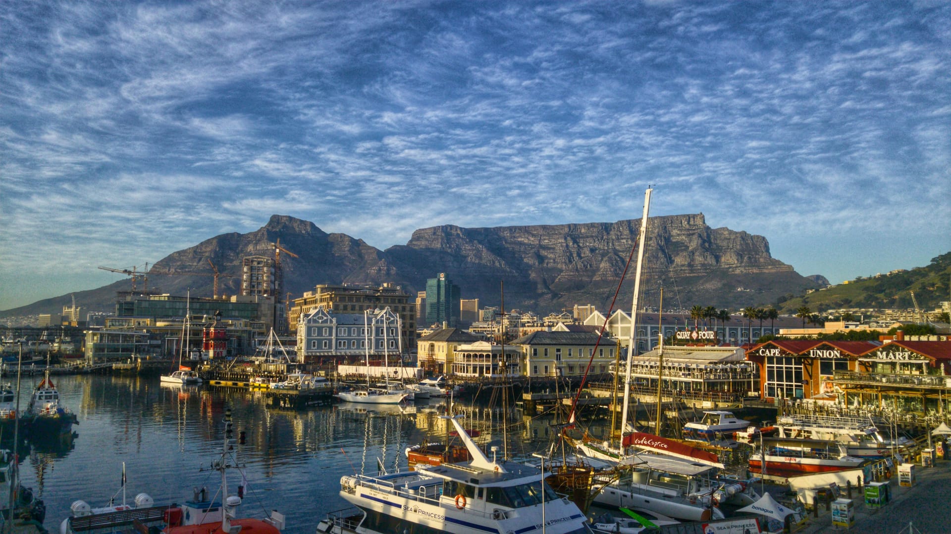 View of the V&A Waterfront harbor with boats in the foreground and Table Mountain looming in the background under a blue sky.