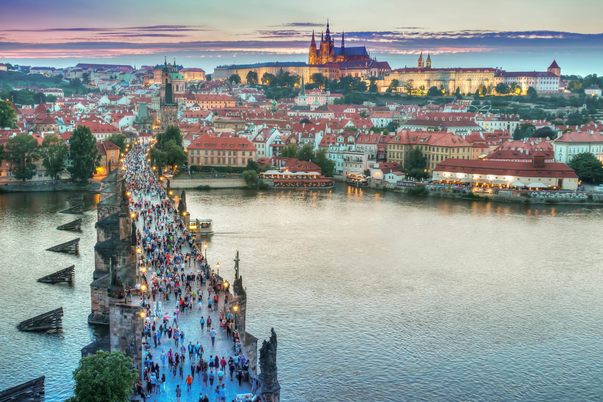 Twilight view of Charles Bridge in Prague with people walking, Vltava River, and Prague Castle illuminated in the background