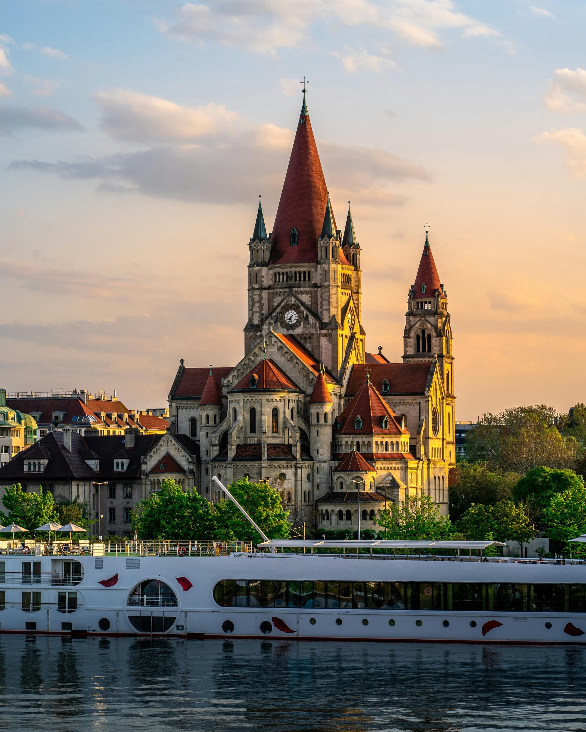 St. Francis of Assisi Church in Vienna with red-tiled spires and a river cruise ship docked along the Danube at sunset