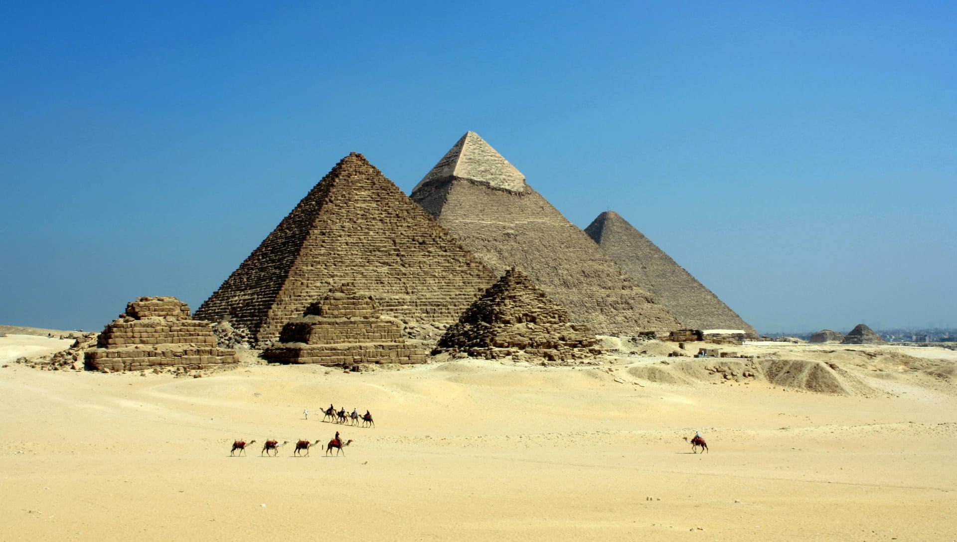 A landscape view of the three Great Pyramids of Giza with a caravan of camels walking across the desert sand in the foreground.