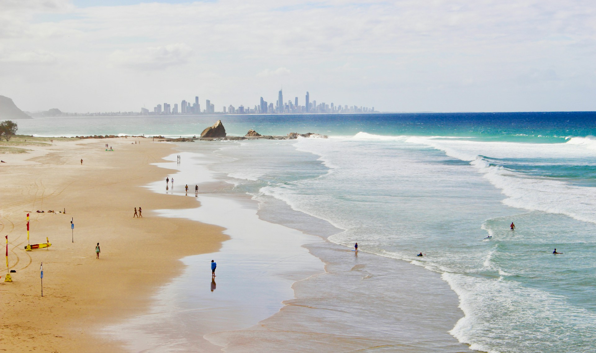 A wide panoramic view of a sandy beach with gentle waves rolling in, and a city skyline visible on the distant horizon under a cloudy sky.