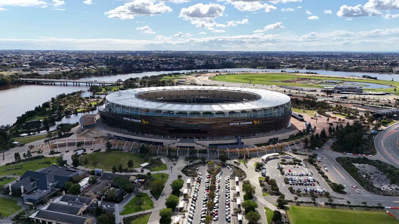 Aerial drone view of the large, circular Optus Stadium and surrounding parklands located next to the river.