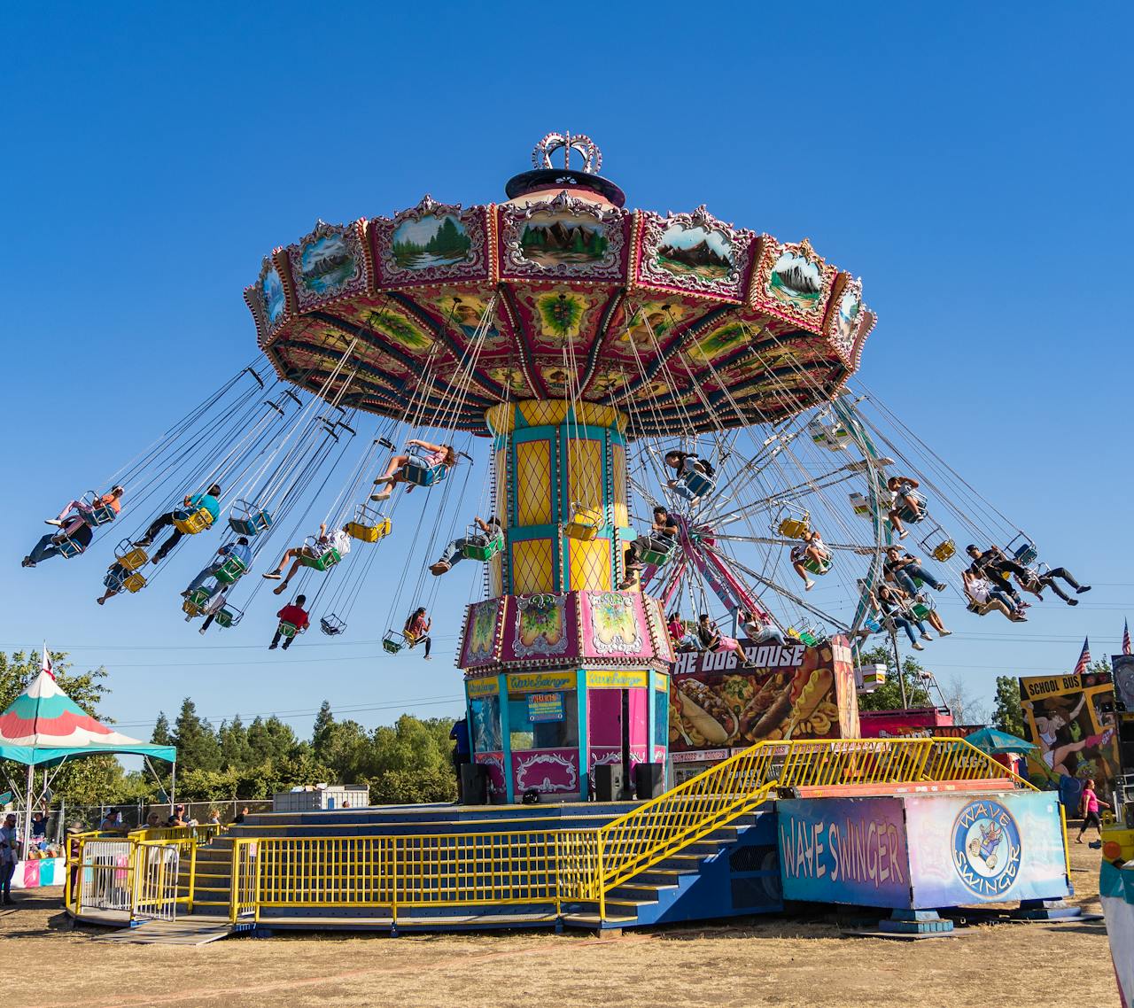 A colorful wave swinger carnival ride spinning with people in the seats against a bright blue sky.