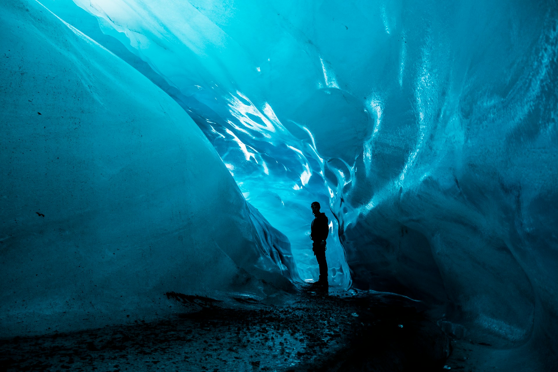 A person standing inside a glacial ice cave, surrounded by smooth, glowing blue ice walls.