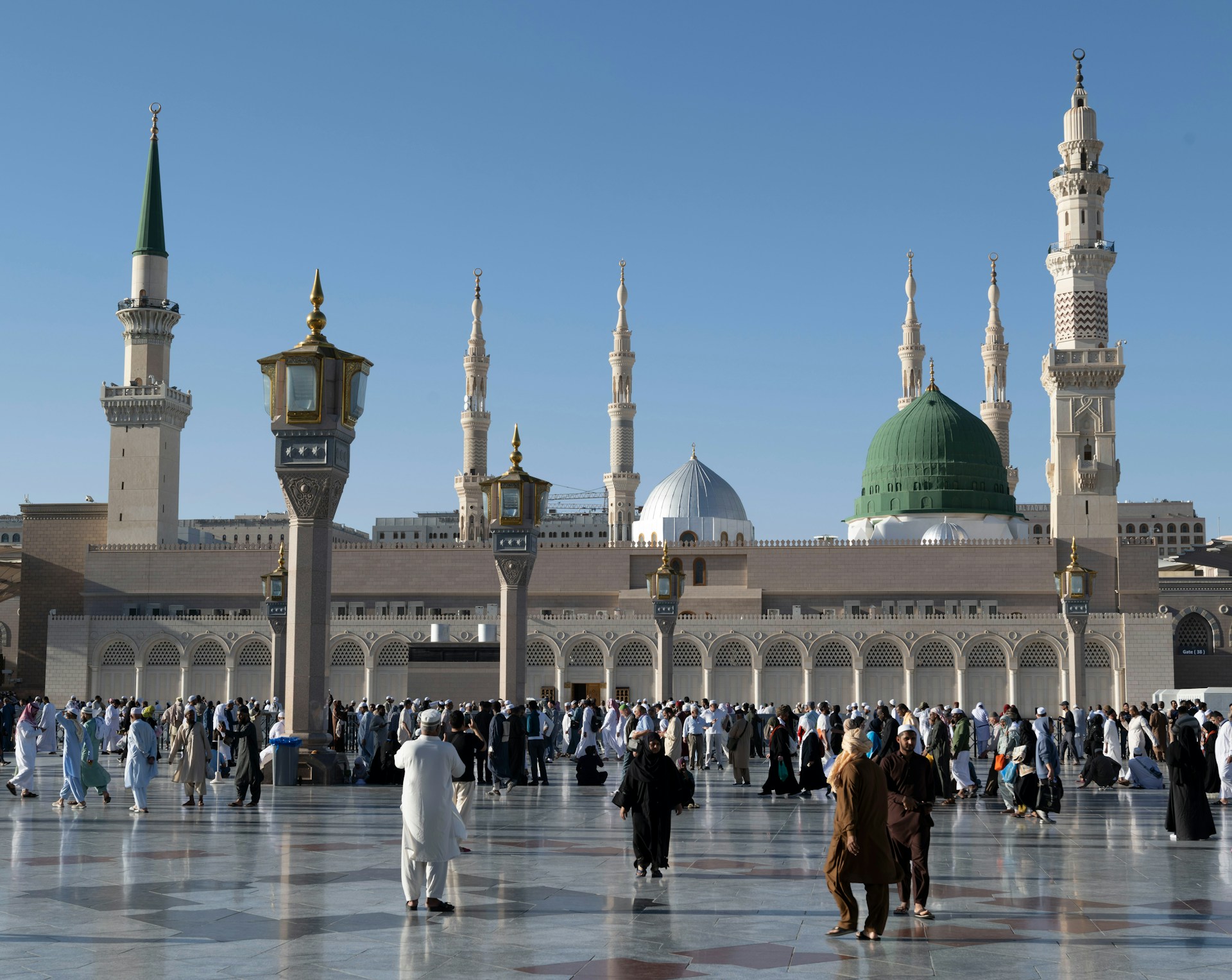 A view of the Green Dome and towering minarets of the Prophet's Mosque in Medina, with people walking in the courtyard.
