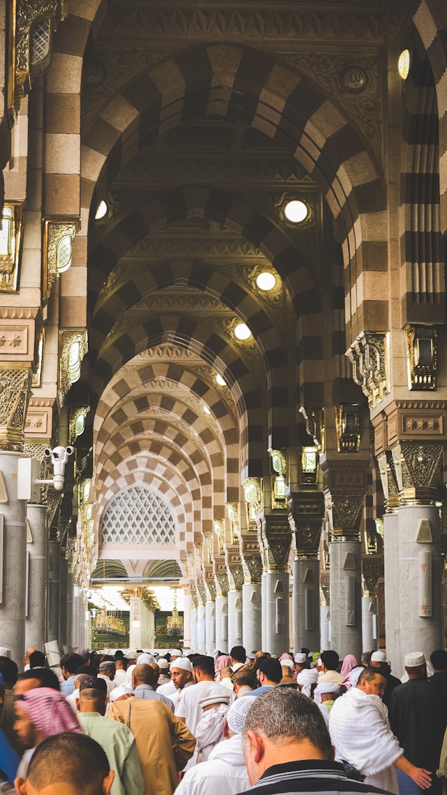 A view down a long hallway of stone columns and arches inside the Prophet's Mosque, filled with worshippers.