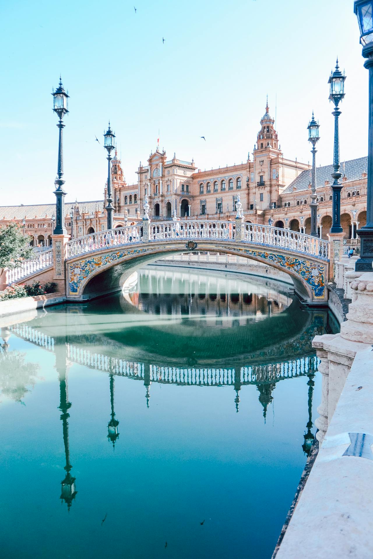 A view of the canal and arched bridges of the Plaza de España in Seville, with the main building's tower reflecting in the water.