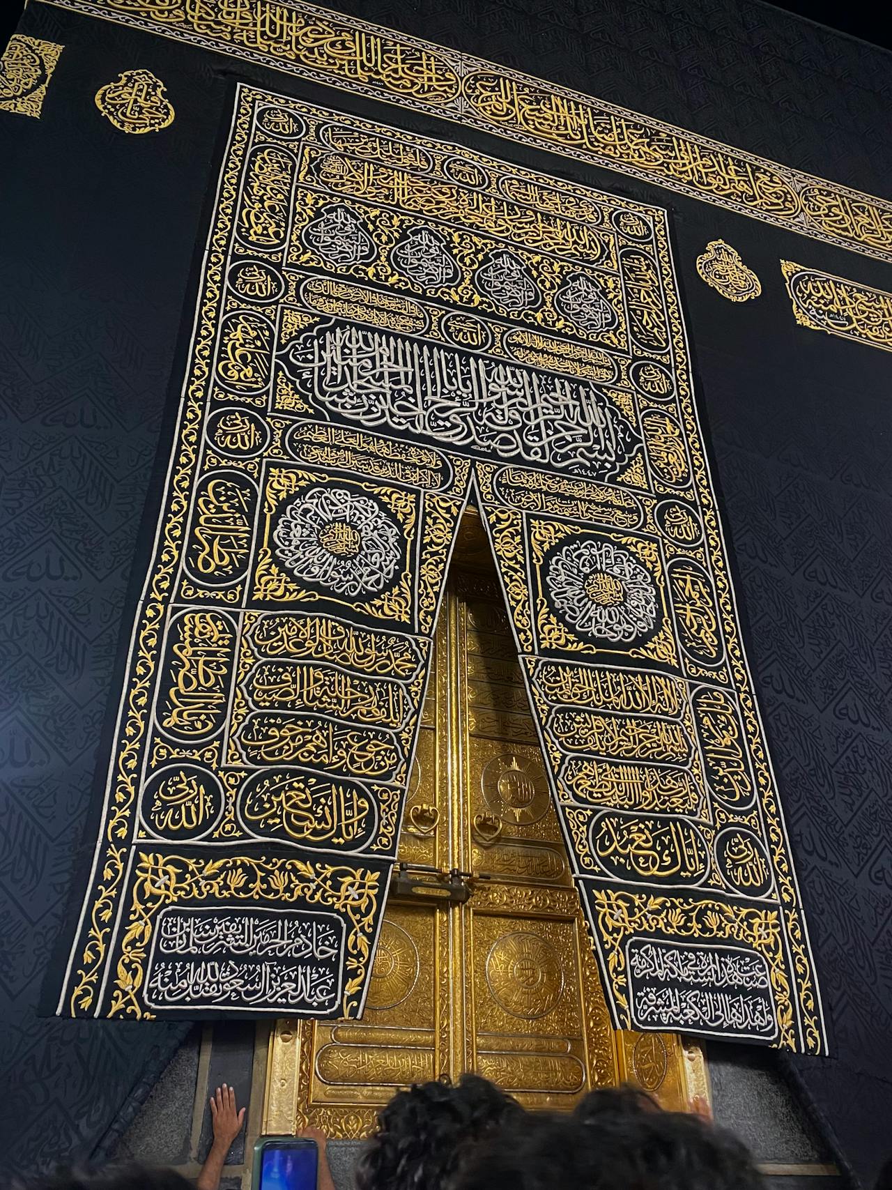 Low-angle close-up view of the golden door of the Kaaba with pilgrims' hands reaching up to touch it.
