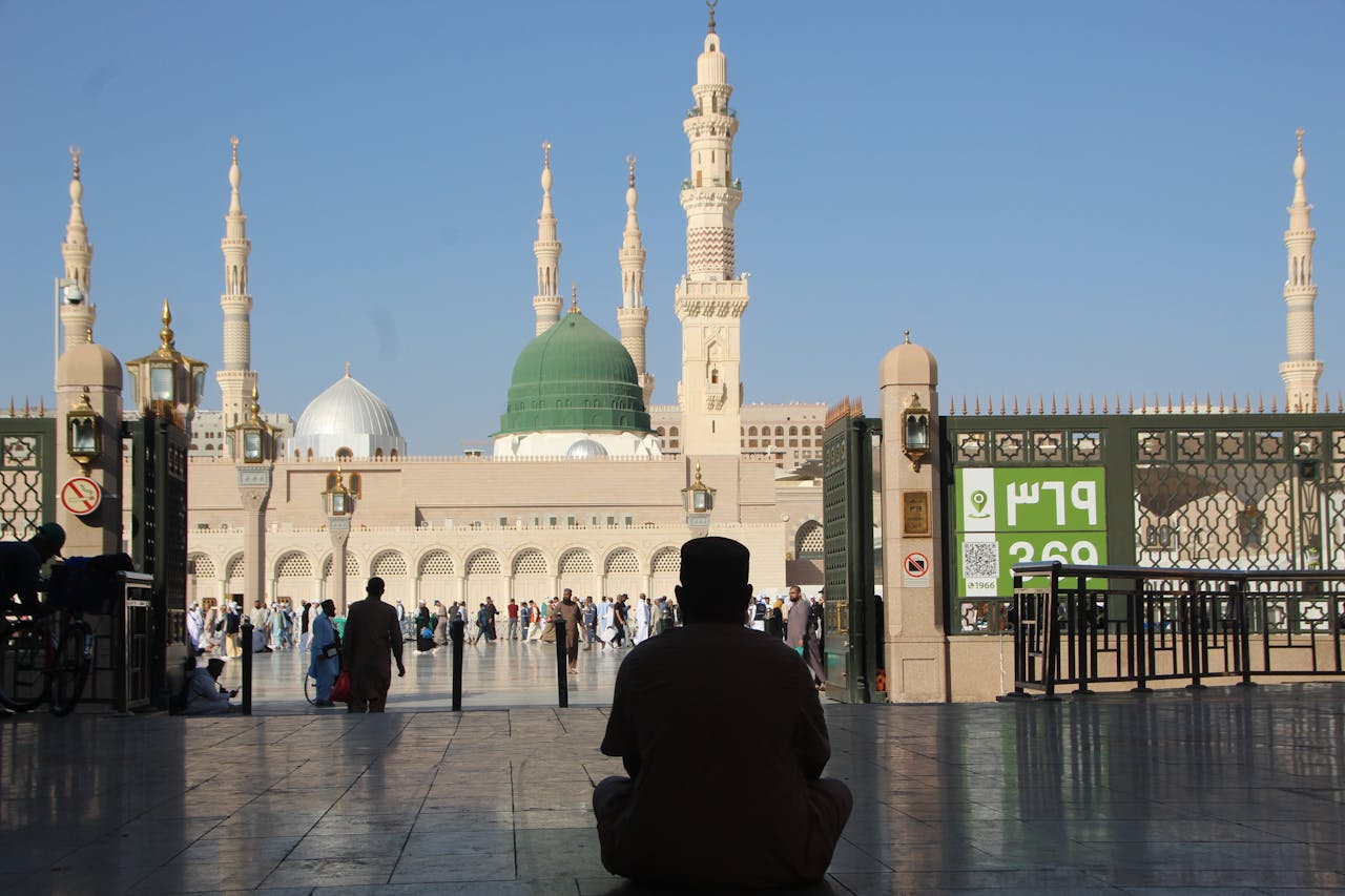 A pilgrim sitting on the ground in the courtyard of Al-Masjid an-Nabawi, looking towards the Green Dome and minarets.
